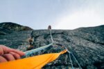 This image is taken from the perspective of a person stood at the bottom of a cliff face. Their left hand is grasping a climbing rope attached to a second person who has already scaled the cliff face.