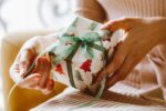 This image shows a person holding a Christmas present wrapped in white paper with green and red trees. The gift is tied with a green taffeta ribbon that the person is pulling to open the gift.