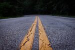 This image is taken from the surface of a grey tarmaced road painted with two parrallel yellow lines along the centre. The camera looks along the yellow lines, which stretch out into the distance.