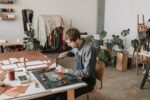 This image shows a man using tools to make a leather product while sitting at a desk in a brightly lit room.