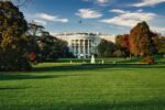 A photo of the US White House taken looking across the lawns. The image is framed by trees and features a water fountain in the centre.